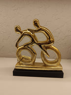 Stylized golden sculpture of two abstract human figures riding bicycles