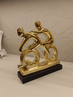 Stylized golden sculpture of two abstract human figures riding bicycles