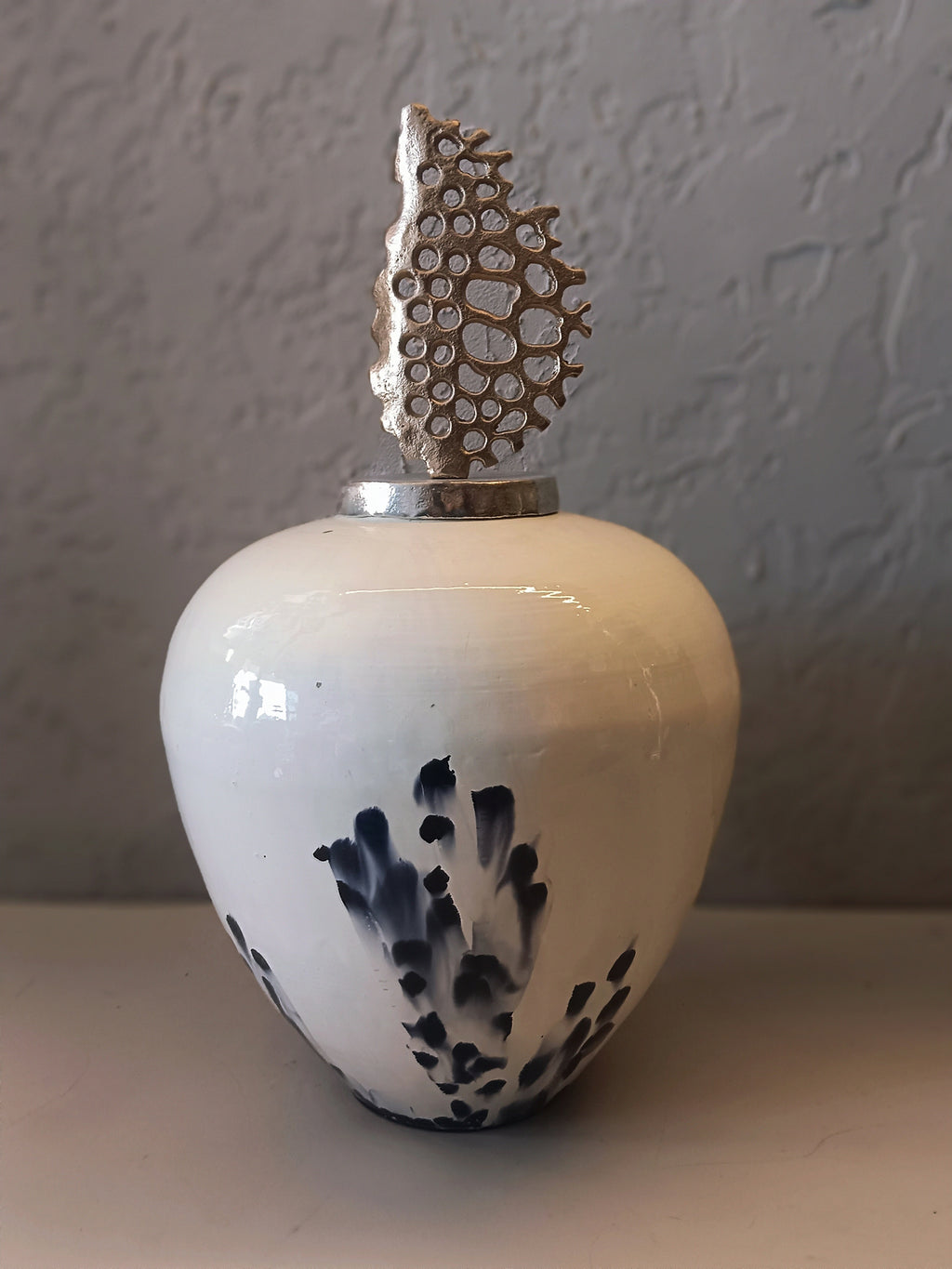 A refined decorative metal vase with a glossy white finish, accented by soft navy brush patterns and crowned with an artistic metallic coral-inspired top